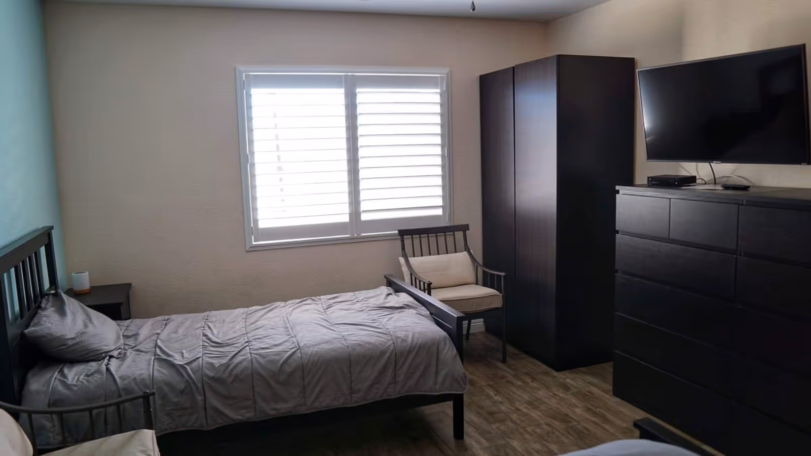 A simple bedroom with a single bed covered in gray bedding, a wooden chair with a cushion, a dark wooden wardrobe, a matching dresser with a flat-screen TV mounted above it, and a window with white blinds letting in natural light.