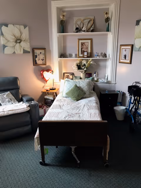 A cozy assisted living bedroom featuring a single bed with white and green pillows, a bedside table with a lamp and heart-shaped decoration, a recliner chair with a blanket, framed pictures and floral artwork on the walls, built-in shelves with decorative items, and a walker near a small black nightstand and wastebasket.
