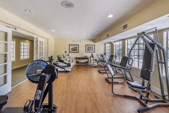 A fitness room with various exercise equipment including stationary bikes, elliptical machines, and weight machines. The room has wooden flooring, large windows with blinds allowing natural light, and framed pictures on the far wall. There is an open doorway leading to another room.