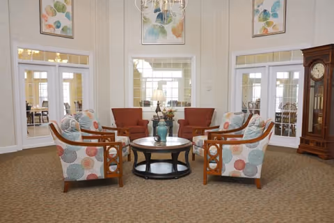A senior living lounge with patterned armchairs arranged around a round coffee table, additional seating, a grandfather clock, and wall art.