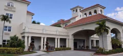 White stucco senior living facility with a red-tiled roof, arches, and palm trees at entrance.