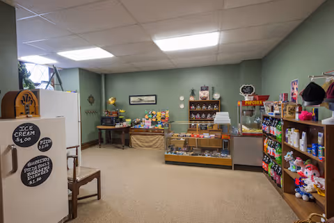 Interior view of a small shop area inside a facility with green walls and beige carpet. There is a white refrigerator on the left with signs advertising ice cream and snacks. Shelves and display cases hold various items including stuffed animals, snacks, and a popcorn machine. The room is lit by ceiling lights and has a window on the left side.