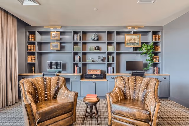 Two brown leather armchairs and a small round table facing blue built-in shelving and desk workspaces in a lounge area.