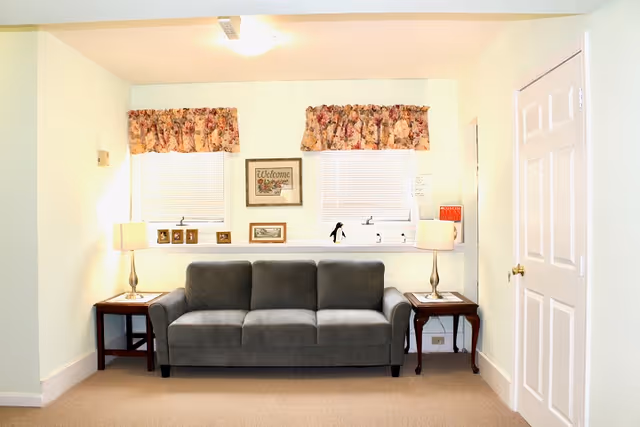 A cozy living room area with a gray three-seater sofa centered against a wall with two windows above it. The windows have floral valances and white blinds. On either side of the sofa are wooden side tables with matching lamps. Various framed pictures and a 'Welcome' sign are displayed on the windowsill behind the sofa. A closed white door is visible on the right side of the image.