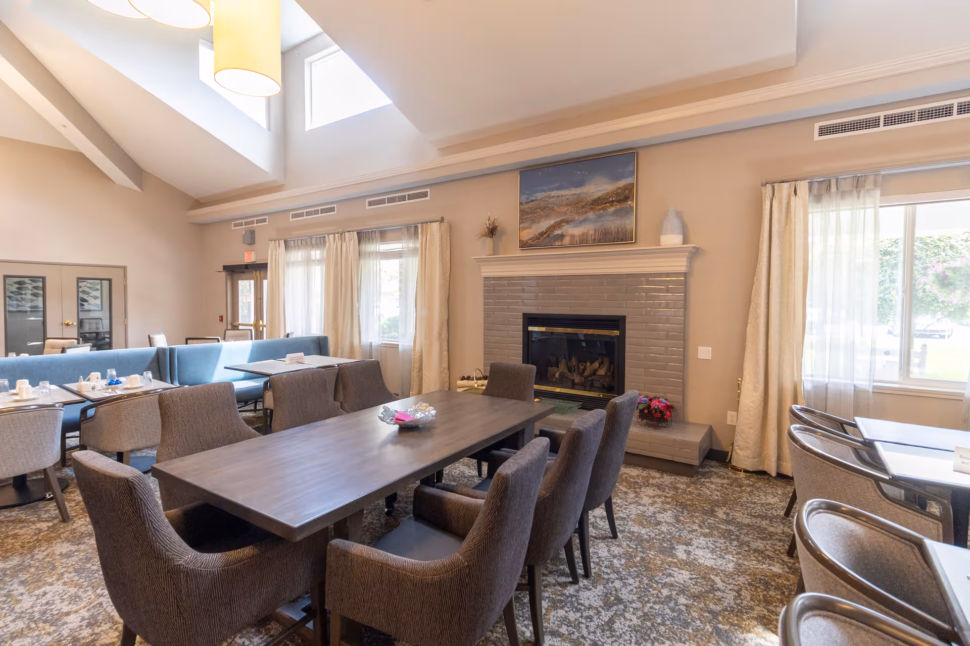 A spacious dining room with a large wooden table surrounded by eight upholstered chairs. The room features a fireplace with a painting above it, large windows with sheer curtains allowing natural light, and additional seating areas with tables and chairs. The ceiling has skylights and hanging pendant lights.