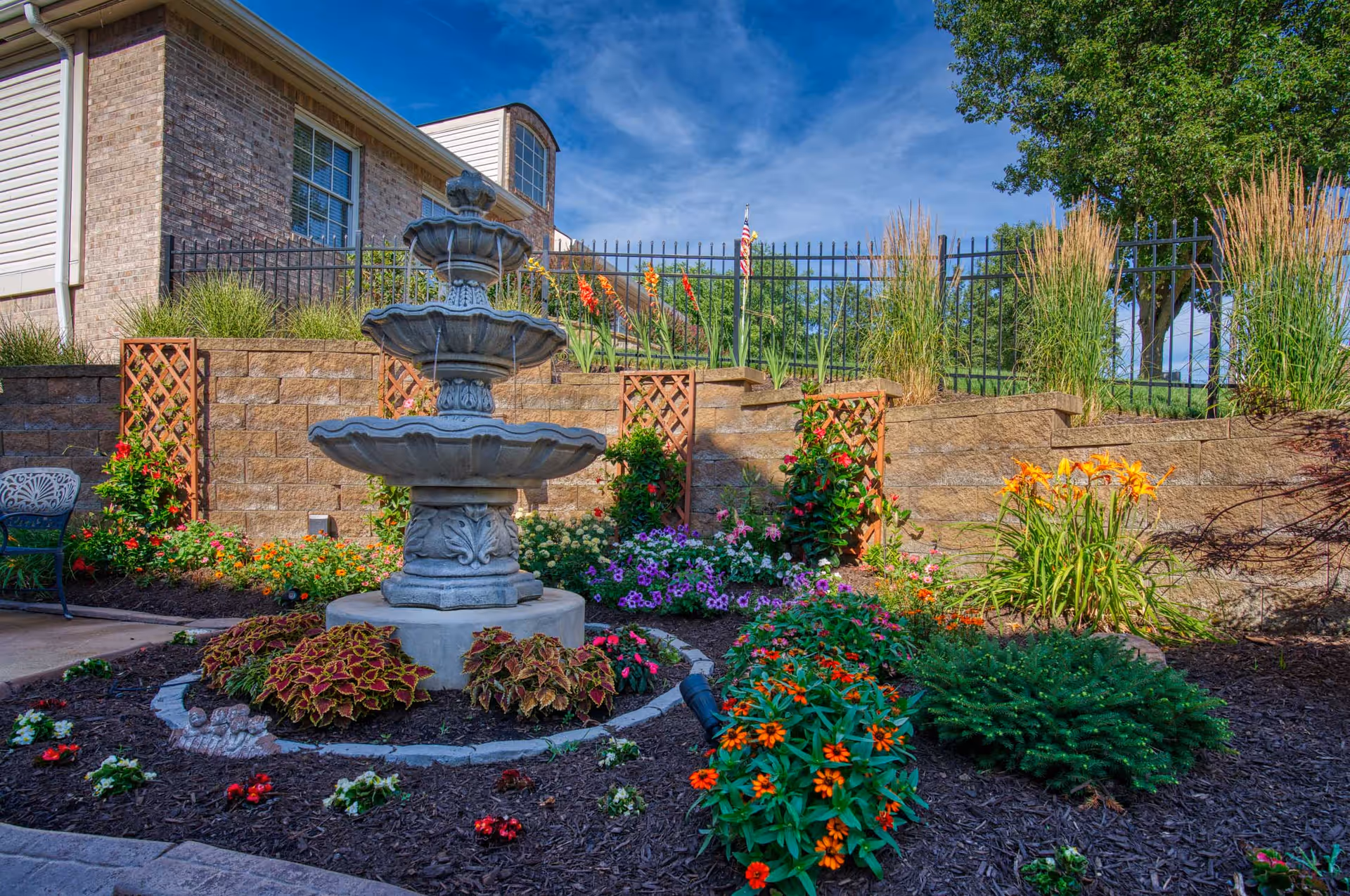 A landscaped outdoor garden area featuring a multi-tiered stone fountain surrounded by colorful flowers and plants. Behind the garden is a retaining wall with lattice panels and tall grasses, with a brick building and a black metal fence in the background under a blue sky.