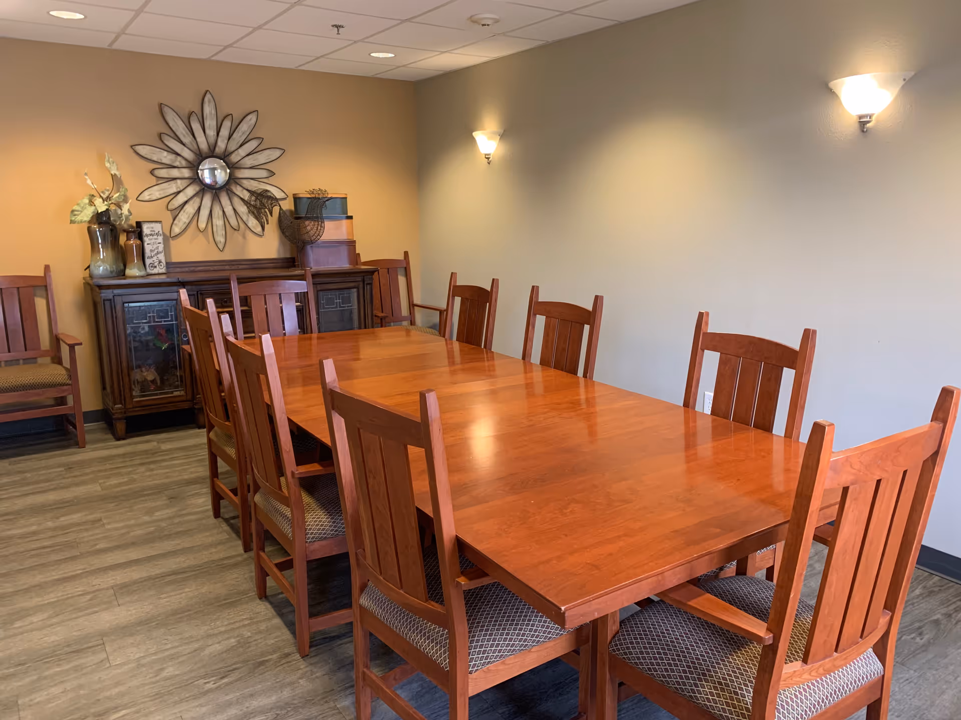 A dining room with a long wooden table surrounded by ten wooden chairs with cushioned seats. The room has light-colored walls with two wall sconces providing light. Against one wall is a wooden cabinet with decorative items on top, including a large flower-shaped mirror and vases.