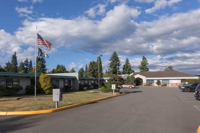 Exterior view of Mountain View Rehabilitation and Care Center showing a single-story building with a parking lot, an American flag and another flag on flagpoles, surrounded by trees and a partly cloudy sky.