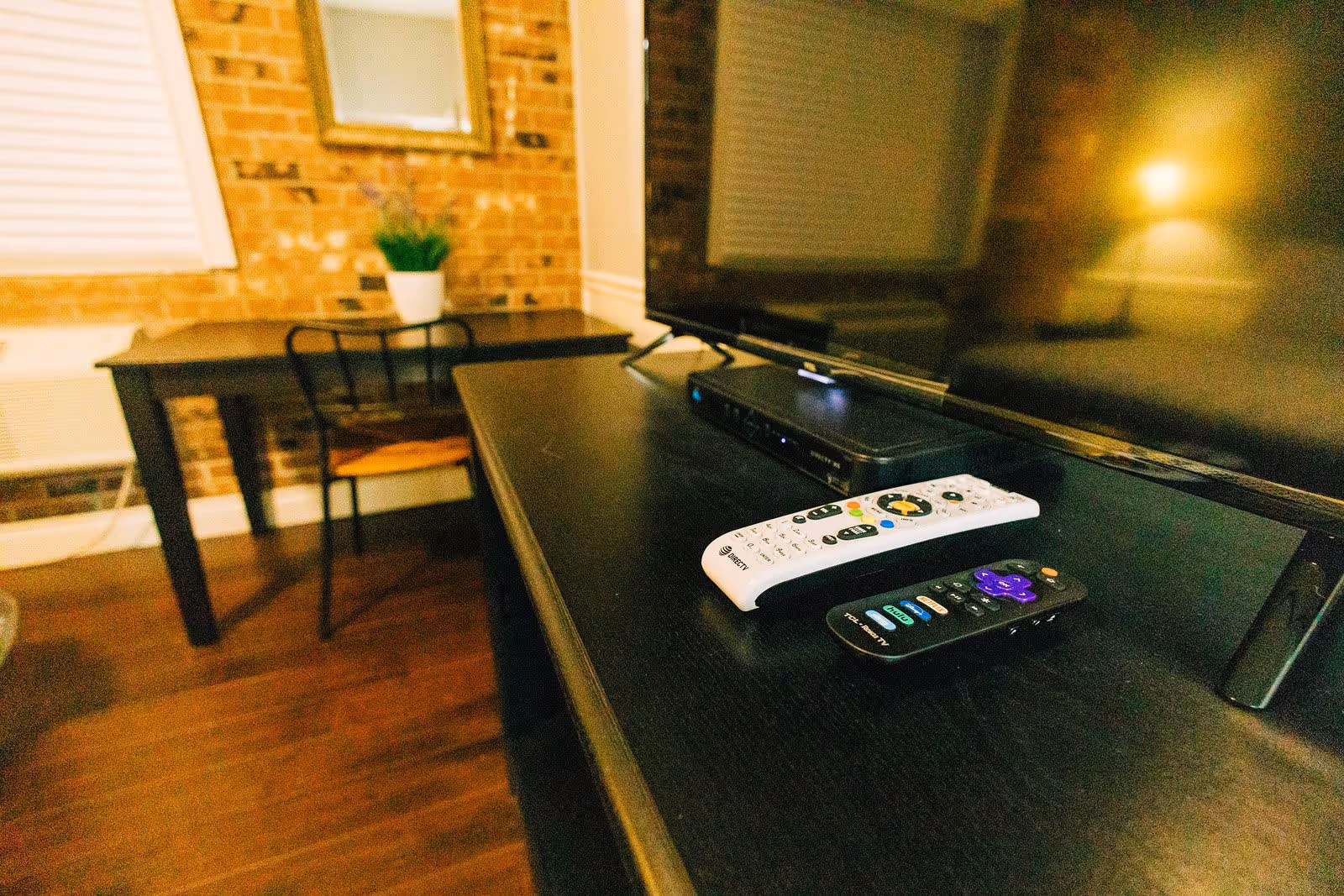 Close-up view of a black TV stand with two remote controls and a cable box in front of a large flat-screen TV. In the background, there is a wooden table with a black metal chair, a potted plant, a mirror on a brick wall, and a window with closed blinds.