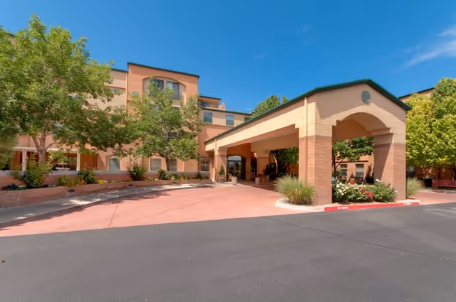 Exterior view of The Woodmark at Uptown senior living facility showing the main entrance with a covered drop-off area, surrounded by trees and landscaping under a clear blue sky.