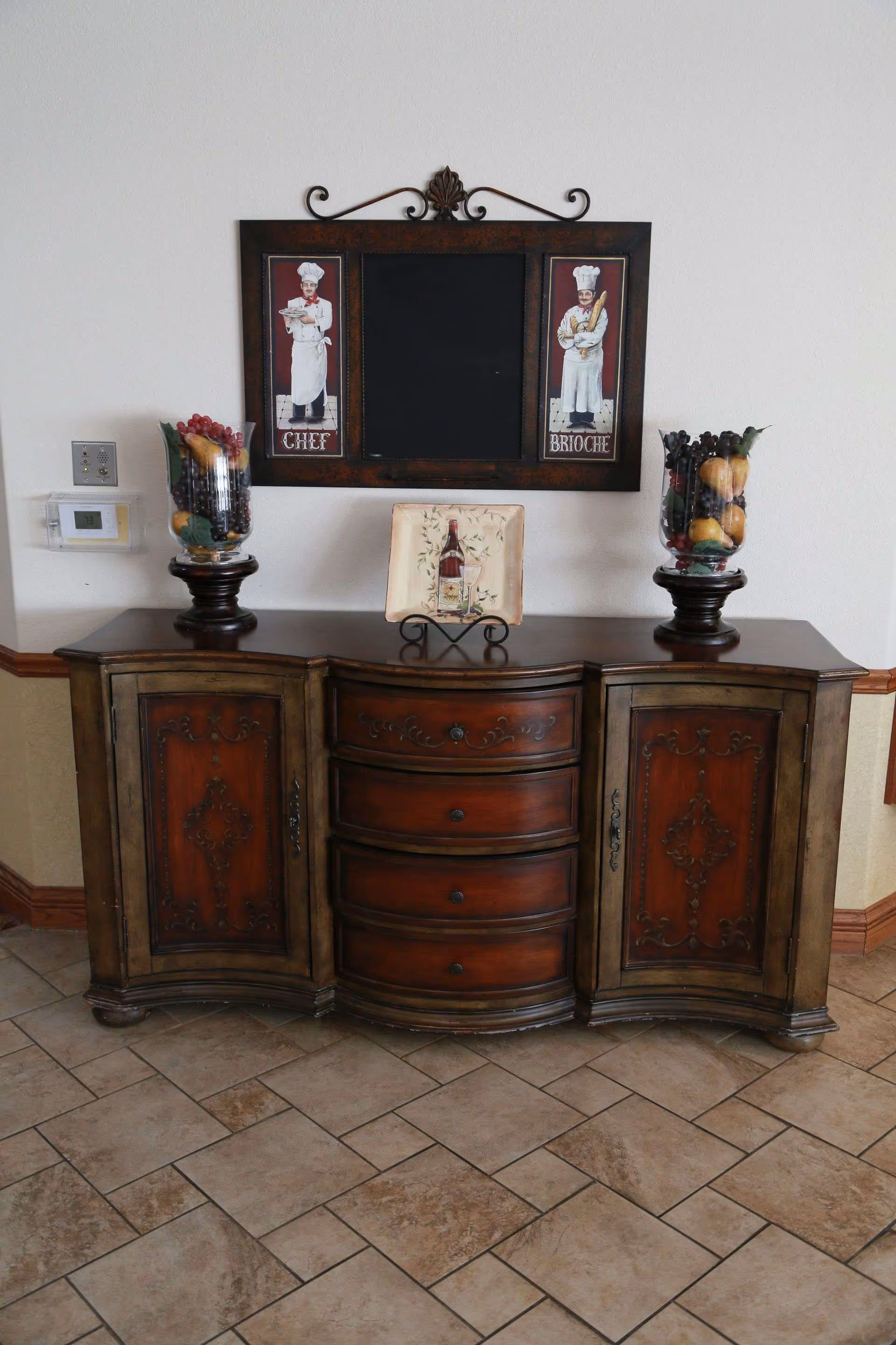 Ornate wooden console table with decorative vases, a plate display and framed artwork on a tiled floor against a white wall.