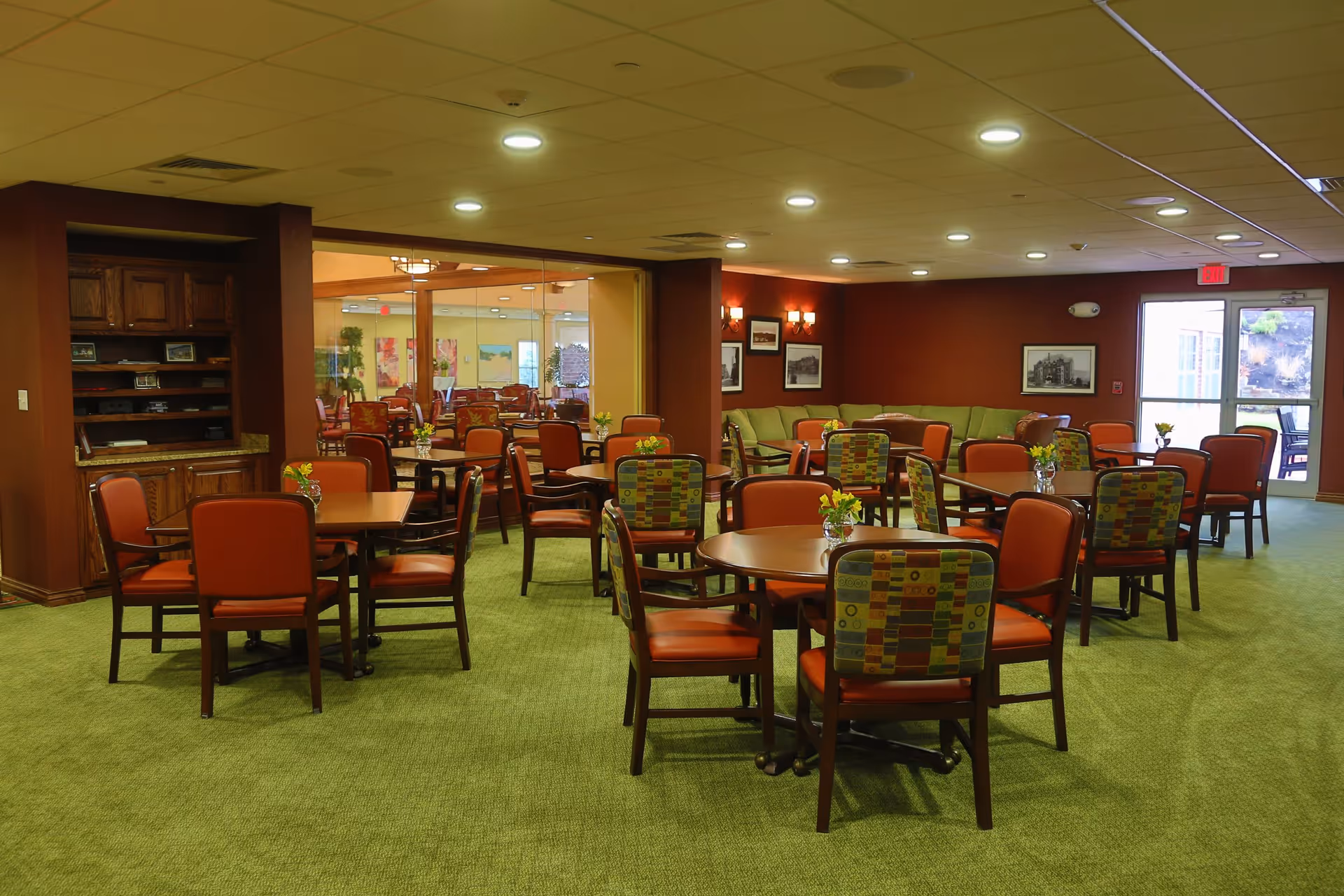 A spacious dining area in a retirement resort with multiple round tables surrounded by chairs. The chairs have orange seats and backs with some featuring a colorful patterned fabric. The room has green carpet flooring, warm red walls, and soft ceiling lighting. There is a glass partition separating this area from another similar dining space, and a green cushioned seating area is visible against the far wall. Small flower arrangements are placed on each table.