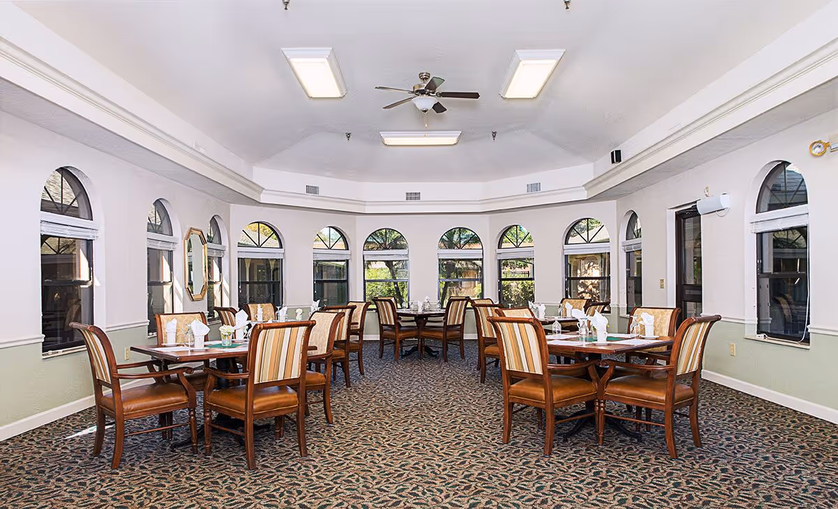 A bright dining room with multiple wooden tables and chairs arranged neatly. Each table is set with napkins and glassware. The room features large arched windows allowing natural light to enter, a ceiling fan, and overhead fluorescent lights. The carpet has a patterned design, and the walls are painted in light colors with a green lower half and white upper half.