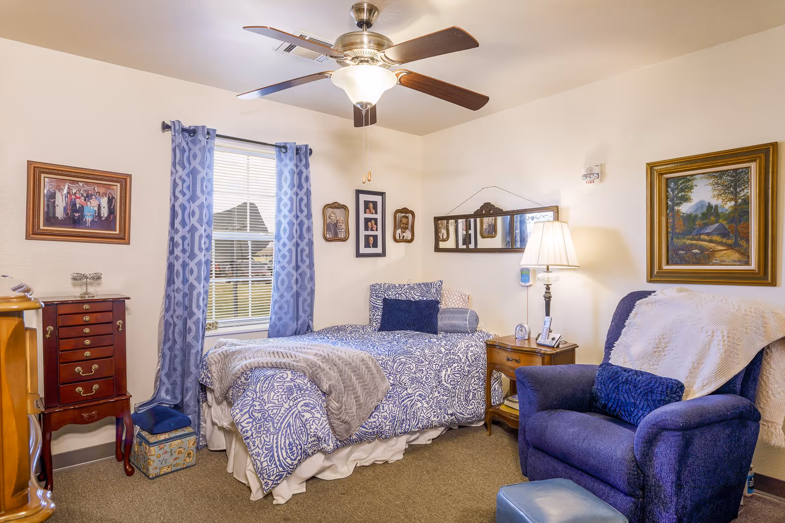 Cozy furnished bedroom with a single bed, blue armchair, nightstand, ceiling fan and framed wall art.
