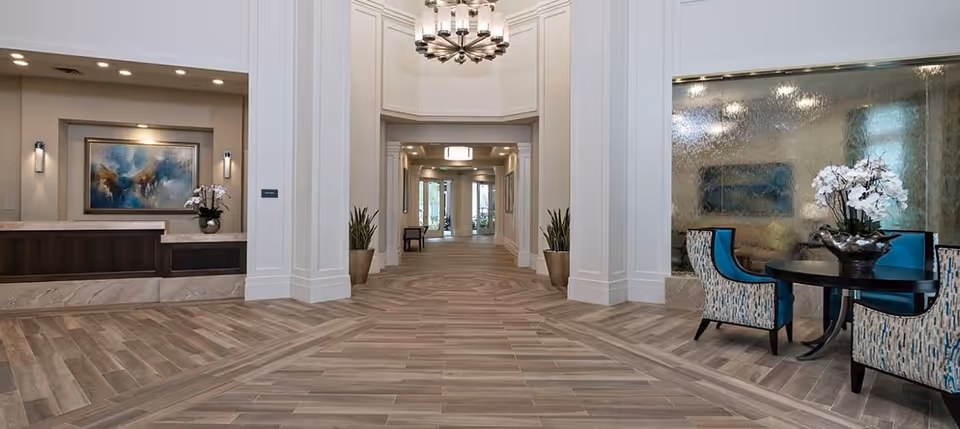 Spacious and elegant interior hallway of a senior living facility with wood-patterned flooring, white paneled walls, a modern chandelier, a reception desk with a painting and orchid, and a seating area with patterned chairs and a round table with a flower arrangement.