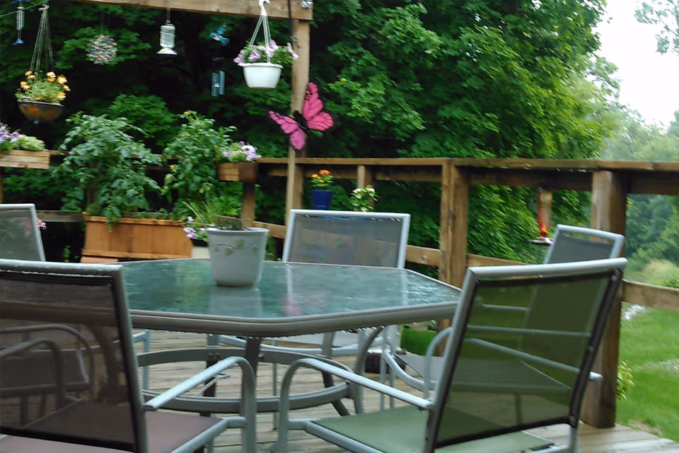 Outdoor wooden deck with a glass-top table surrounded by six metal and mesh chairs. The deck is decorated with potted plants, hanging planters, and a large pink butterfly decoration. Lush green trees and grass are visible in the background.