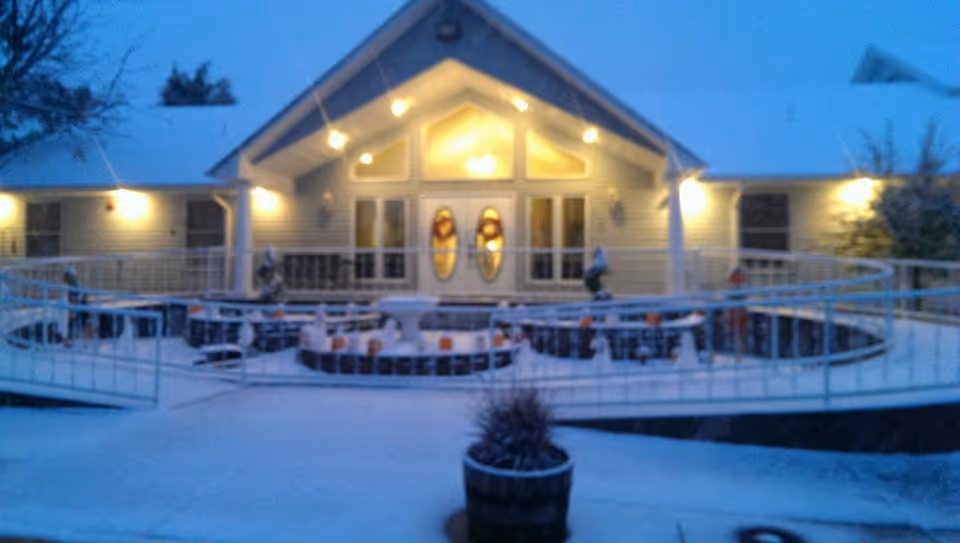 Exterior view of a building at dusk with lights on, featuring a ramp and a circular seating area decorated with pumpkins and small scarecrow figures, with a potted plant in the foreground.