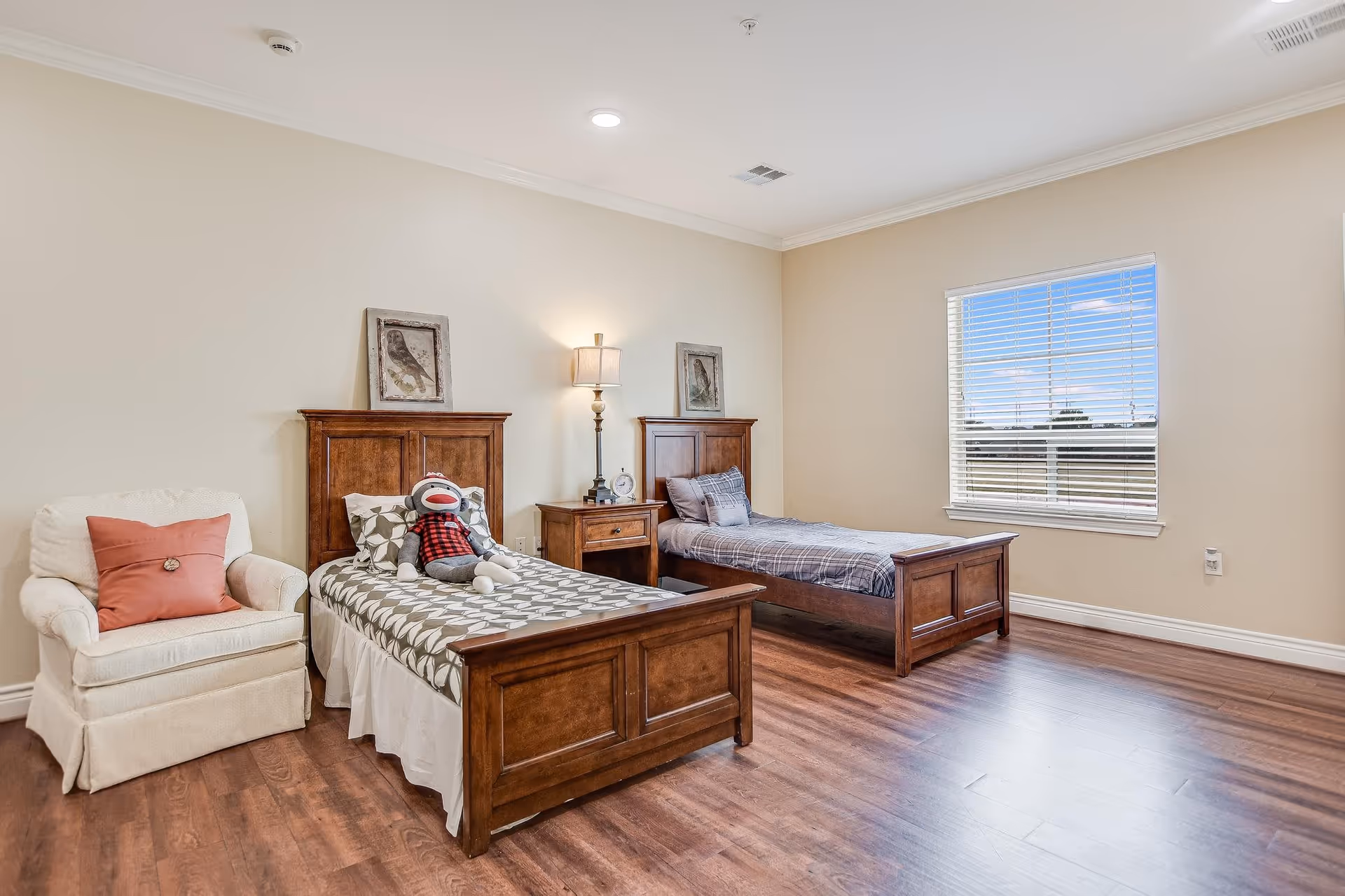 A bright bedroom with two wooden twin beds (one with a stuffed sock monkey), a nightstand and lamp between them, an armchair with a coral pillow, hardwood floors and a window.