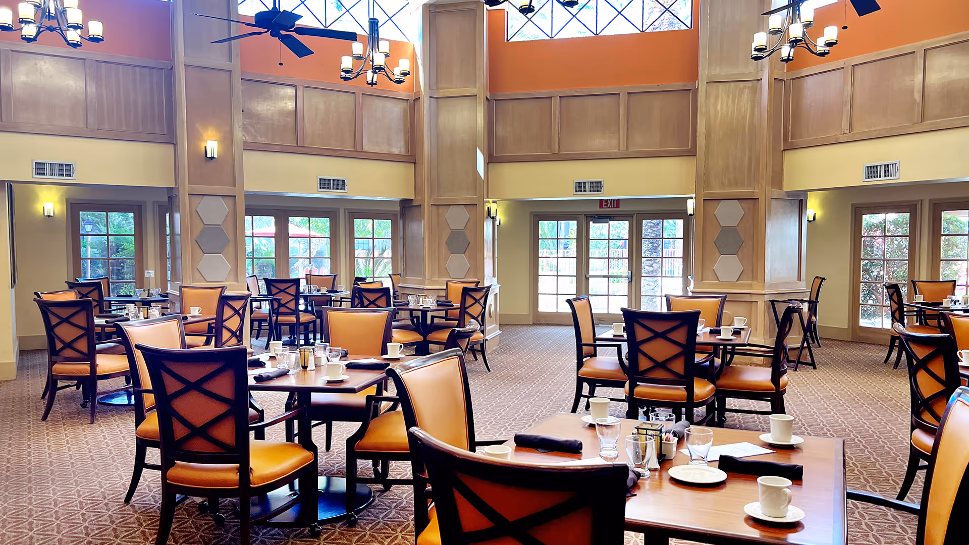 Spacious dining room with multiple round tables and chairs arranged neatly. Each table is set with cups, plates, and napkins. The room features high ceilings with large windows allowing natural light to fill the space, ceiling fans, and elegant chandeliers. The walls have wood paneling and warm tones, creating a welcoming atmosphere.