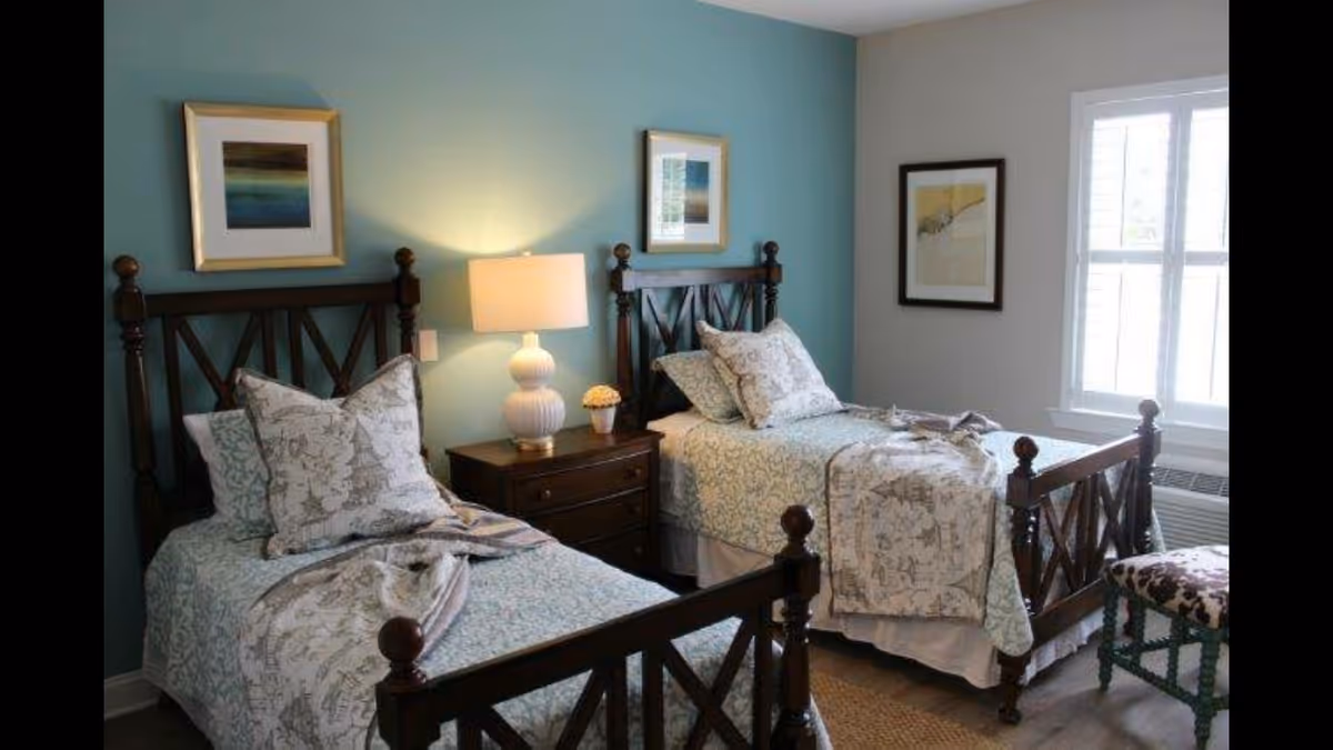 A cozy bedroom with two twin beds featuring dark wooden frames and light patterned bedding. Between the beds is a wooden nightstand with a white lamp and a small potted plant. The walls are painted in two tones, teal behind the beds and light gray on the adjacent wall, which has a window with white blinds and a framed picture. A small upholstered bench with a floral pattern is placed near the window.