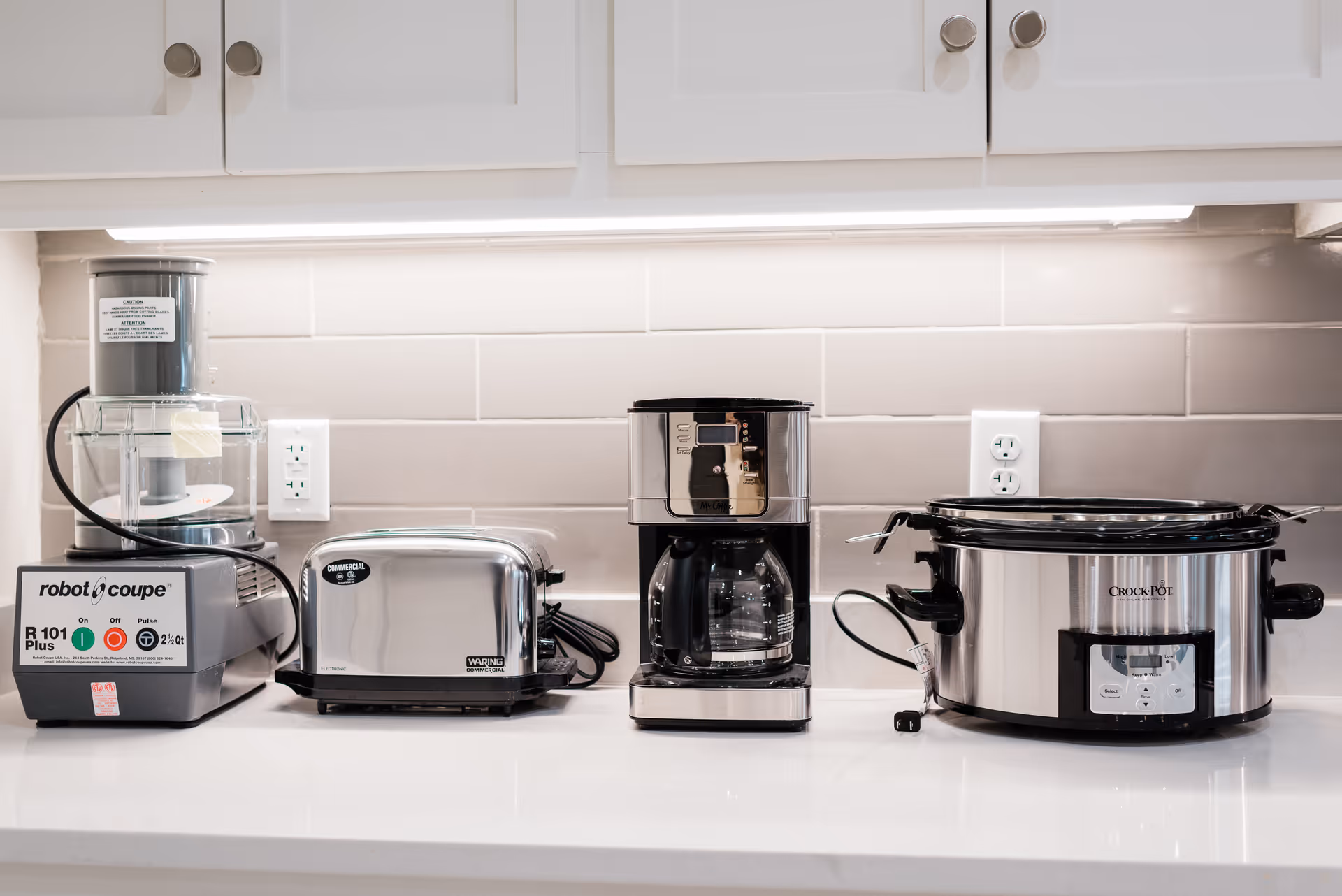 A kitchen countertop with four stainless steel appliances lined up: a Robot Coupe food processor, a two-slice toaster, a drip coffee maker, and a Crock-Pot slow cooker. The background features white subway tile backsplash and white cabinets with silver knobs.