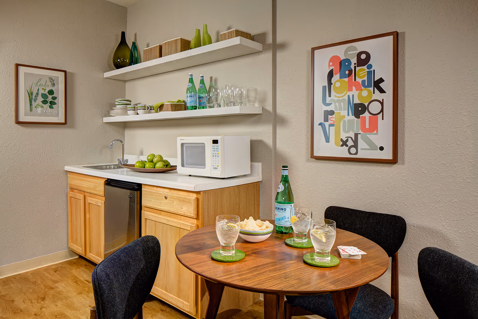 A small kitchenette area with wooden cabinets, a white countertop, a sink, a microwave, and two shelves holding glasses, bottles, and baskets. In front of the kitchenette is a round wooden table with three glasses of water with lemon slices, a bowl of chips, a bottle of sparkling water, and a deck of cards. The room has light-colored walls with two framed artworks, one with herbs and the other with colorful abstract letters. Three dark upholstered chairs surround the table.