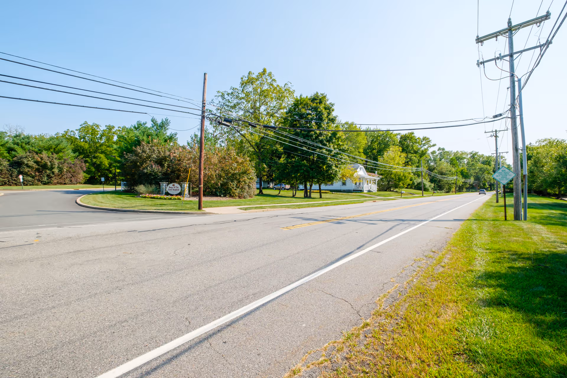 Two-lane road with utility poles and power lines running past grassy lawns and trees with a white house set back from the street.