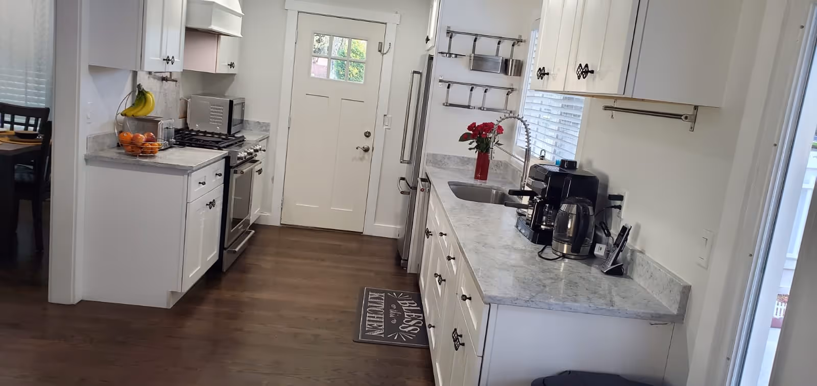 A bright, modern kitchen with white cabinets and marble countertops. The kitchen features a stainless steel stove, microwave, and refrigerator. There is a sink with a tall faucet, a coffee maker, and a kettle on the counter. A small vase with red flowers is placed near the sink. A door with a window is at the end of the kitchen, and a dining area with a table and chairs is visible through an open doorway on the left. The floor is wooden, and there is a mat in front of the sink with the words 'Bless this home'.