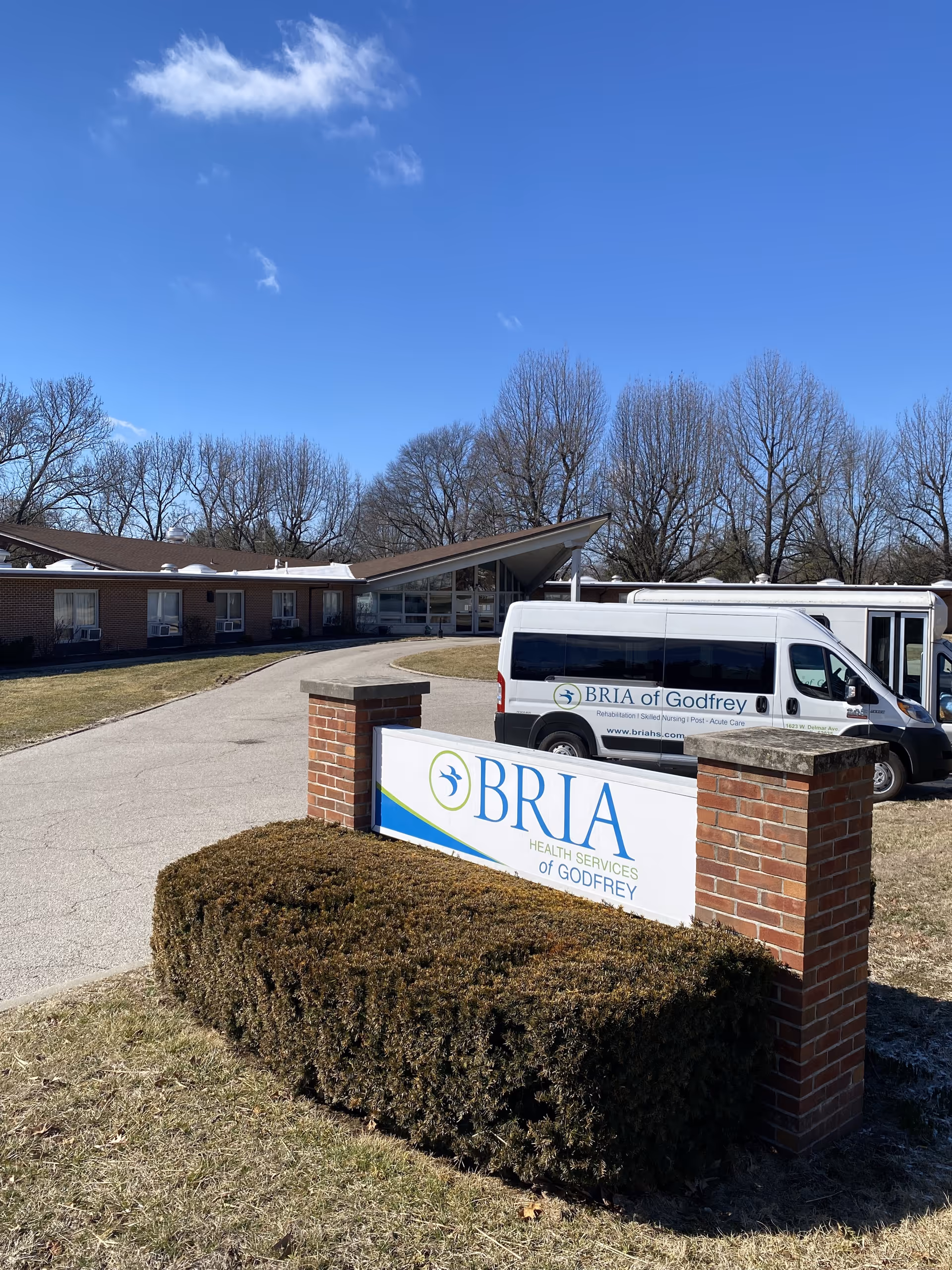 Exterior view of BRIA Health Services of Godfrey building with a clear blue sky. A white van with BRIA of Godfrey branding is parked near the entrance. The facility sign is displayed in front, surrounded by trimmed bushes and a brick structure.