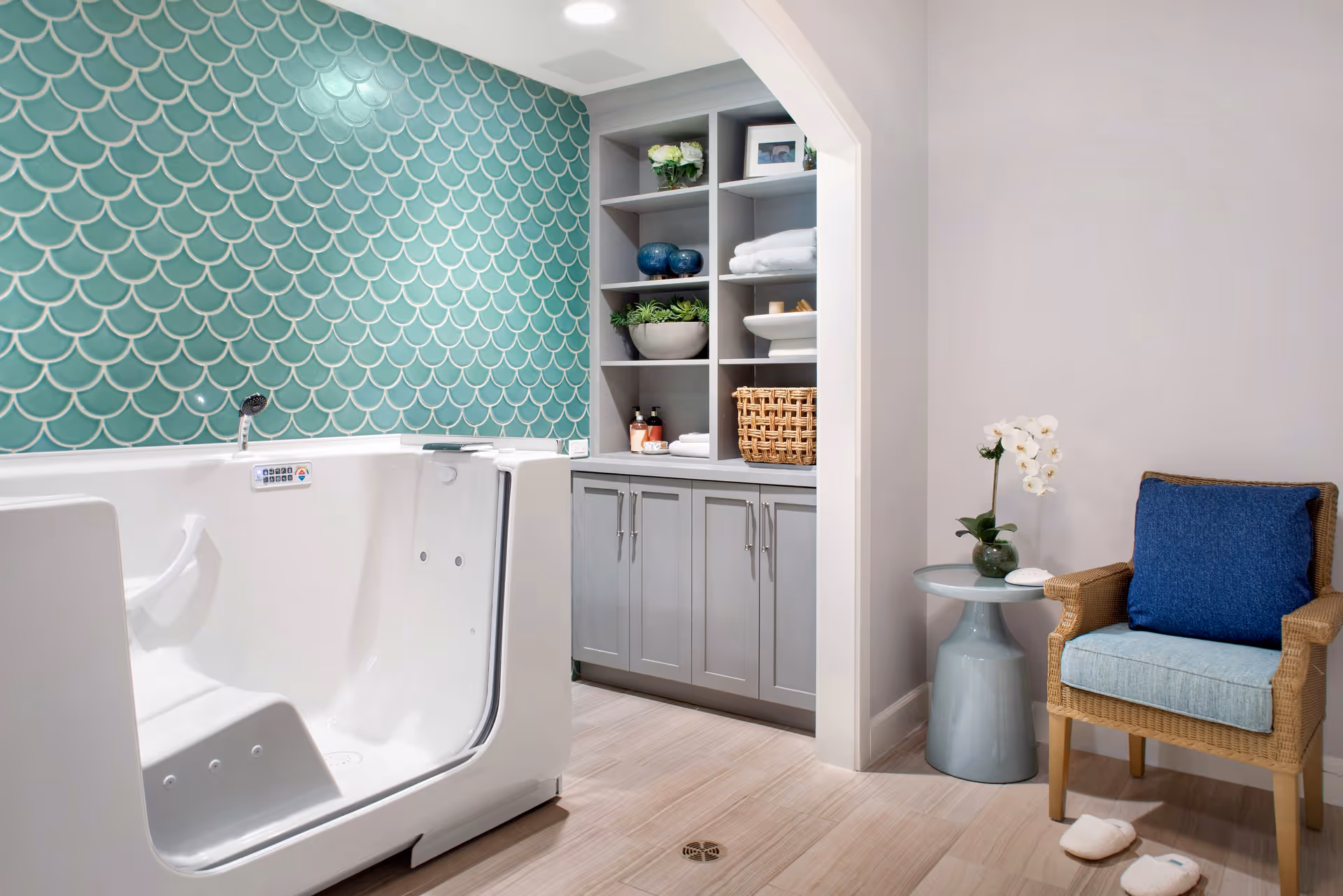 A modern bathroom featuring a white walk-in bathtub with a control panel, a teal fish scale patterned tiled wall, gray built-in shelves with towels, plants, and decorative items, a wicker chair with blue cushions, a small round side table with a white orchid, and slippers on the wooden floor.