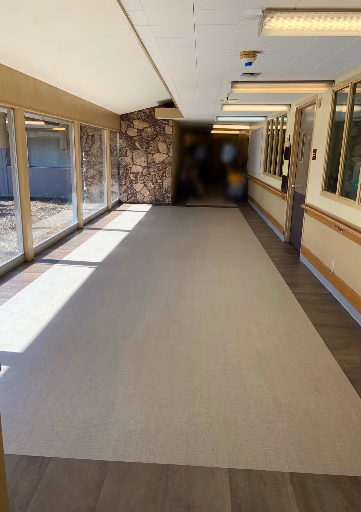 A long, empty hallway in a senior living facility with large windows on the left side letting in natural light. The right side has a handrail and several doors and windows. The far end of the hallway has a stone accent wall.
