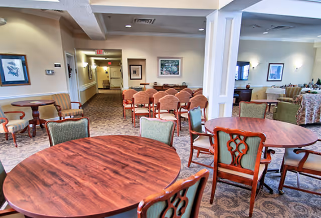 Bright communal dining and lounge area with round wooden tables, upholstered chairs, and seating arranged in an open floor plan.