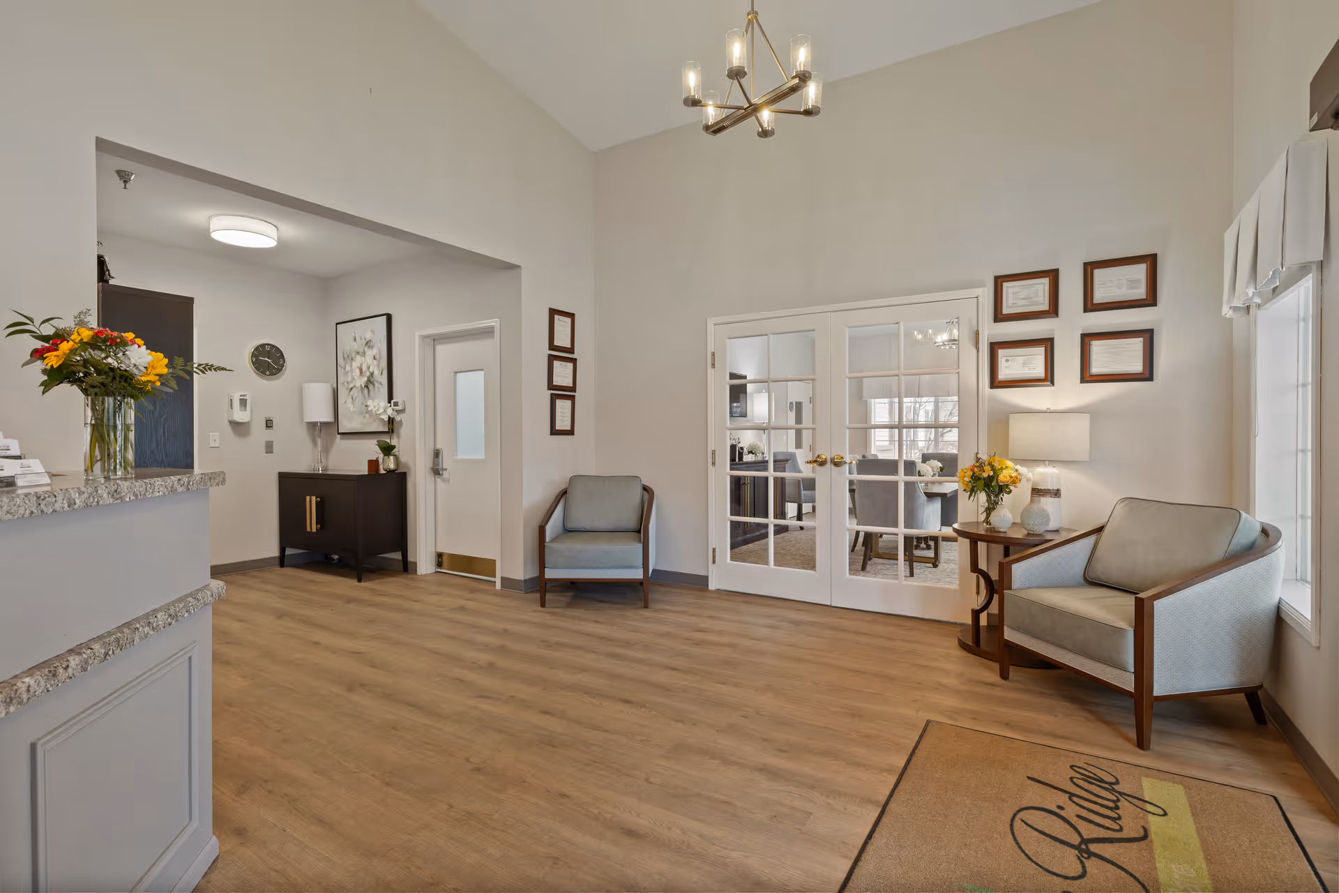 Reception lounge of a memory care facility with chairs, a reception desk with flowers, double glass doors, and a chandelier.