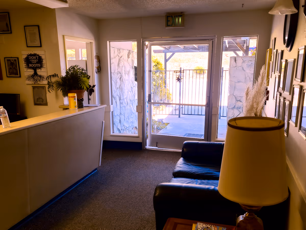 Small assisted-living lobby with a reception desk, leather couch, table lamp and a glass exit door with framed photos on the wall.