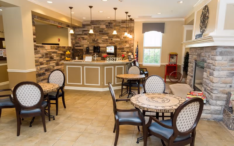 A cozy bistro area inside a senior living facility with round tables and cushioned chairs. The space features a stone accent wall behind a counter labeled 'BISTRO,' pendant lighting, a window with a valance, and a stone fireplace with decorative items nearby.