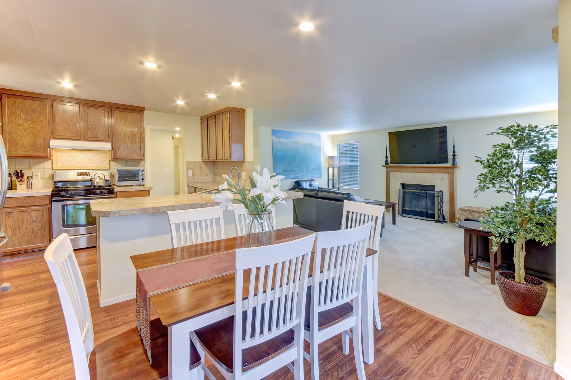 Open concept interior showing a dining area with a wooden table and six white chairs, a kitchen with wooden cabinets and stainless steel appliances, and a living room with a fireplace, mounted TV, black leather sofas, a floor lamp, and a large potted plant.