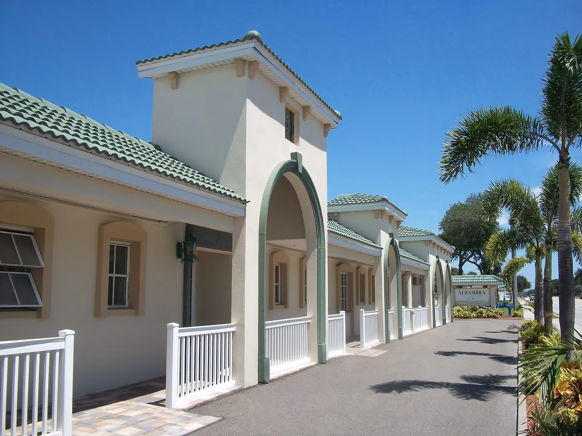 Front exterior of a light-colored Mediterranean-style building with green tile roofs, arched entrances, a driveway, and palm trees.