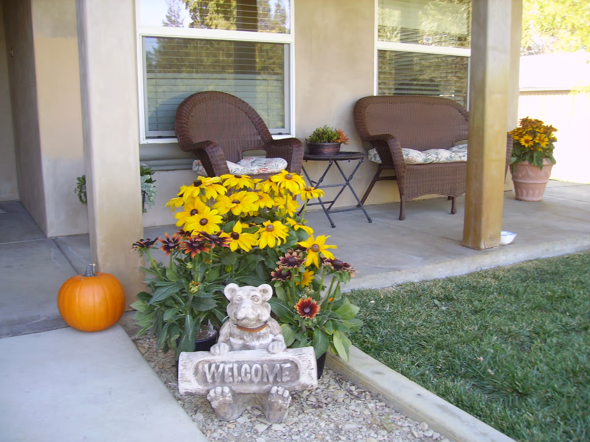 A covered porch area with two wicker chairs with floral cushions, a small black metal table, and potted plants. In front of the porch, there is a garden bed with yellow and dark red flowers and a small stone statue of a bear holding a 'WELCOME' sign. A pumpkin is placed on the concrete walkway near the garden bed, and there is a grassy lawn adjacent to the porch.