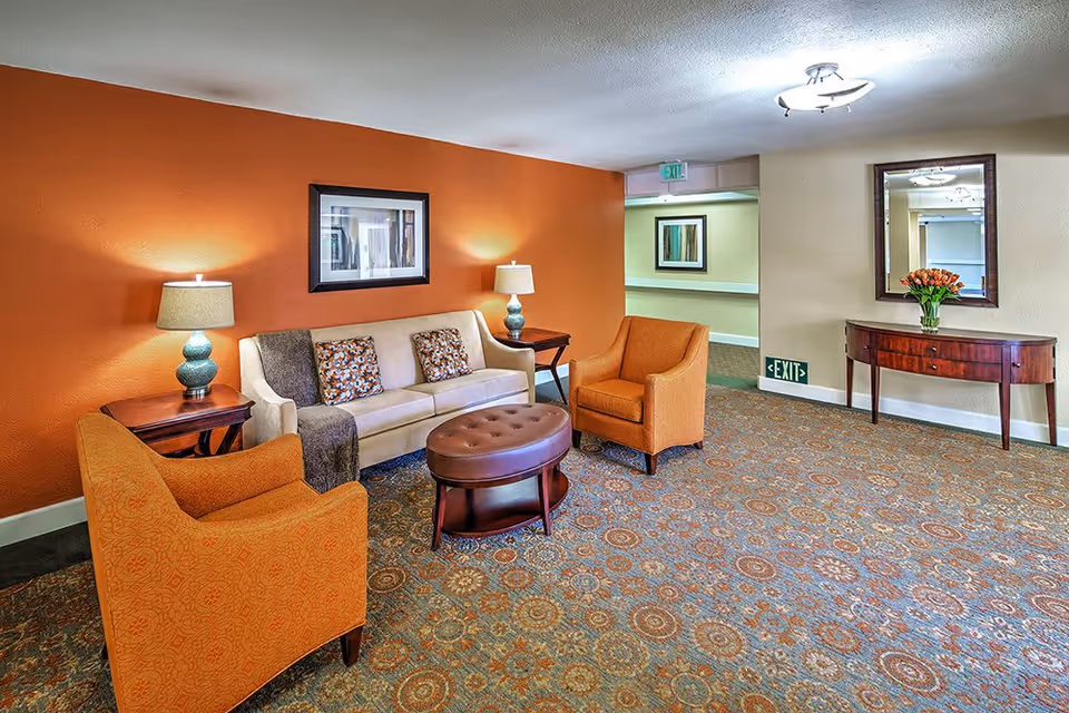 A cozy senior living facility lounge area with an orange accent wall, a beige sofa with patterned cushions, two orange armchairs, a round brown ottoman, two side tables with lamps, a patterned carpet, a wooden console table with a vase of flowers, a large mirror, and an exit sign above a hallway.