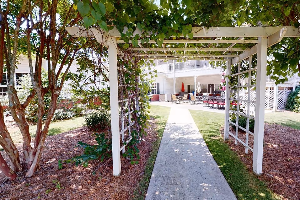 Concrete walkway under a white pergola through landscaped grounds leading to an outdoor patio at a senior living facility.