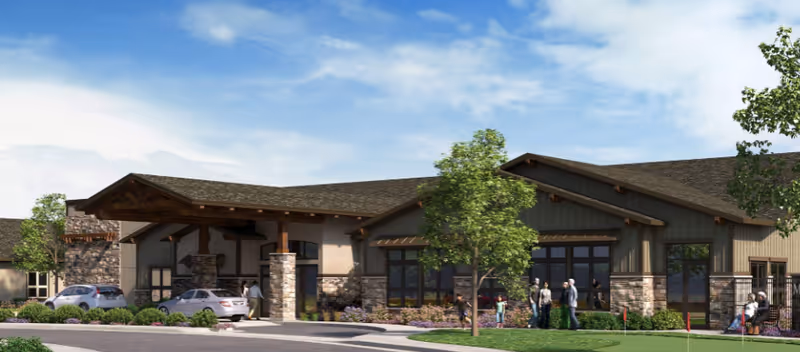 Exterior view of a single-story assisted living memory care facility with stone and wood accents, a covered entrance with cars parked nearby, and people standing and sitting outside on a sunny day with a partly cloudy sky.