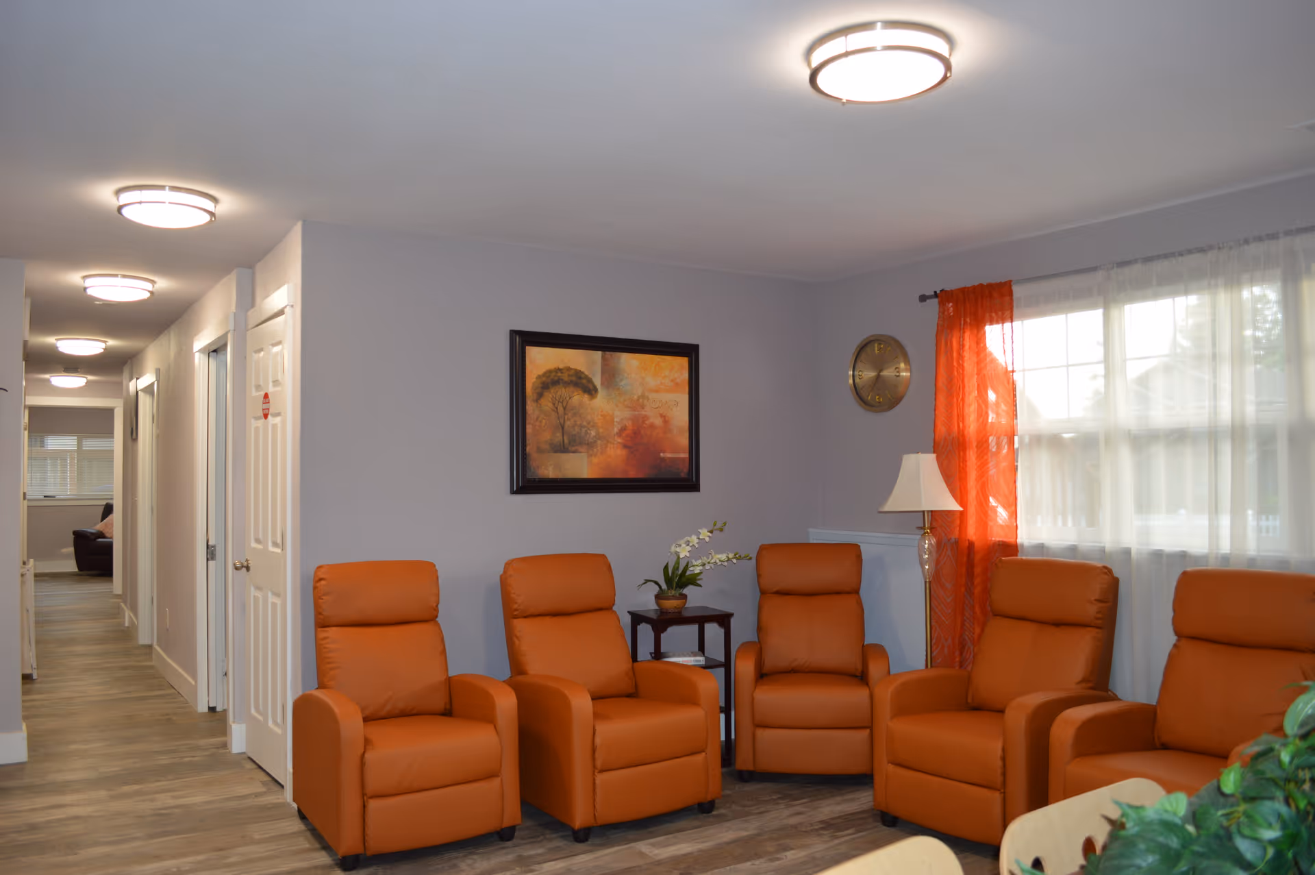 A cozy living room area with five orange recliner chairs arranged in a semi-circle. The room has light gray walls, a wooden floor, a framed painting on the wall, a small side table with a potted plant, a floor lamp, a round wall clock, and a window with sheer white curtains and orange drapes. A hallway with multiple doors is visible to the left.