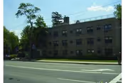 Exterior view of a three-story brick building with multiple windows, surrounded by trees and greenery, located along a street with visible road markings.
