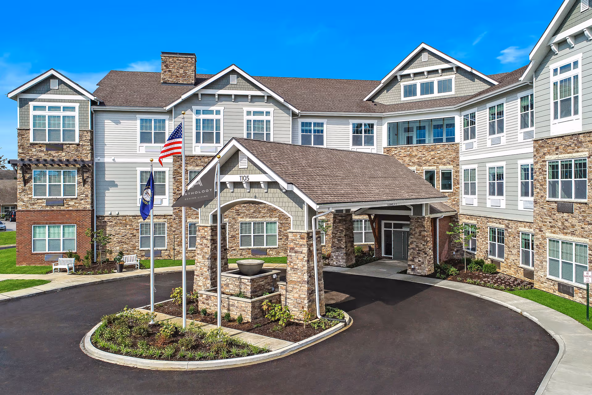 Exterior view of a senior living facility named The Ashton on Dorsey, featuring a large covered entrance with stone pillars, multiple windows, and three flagpoles with flags in front of the building under a clear blue sky.