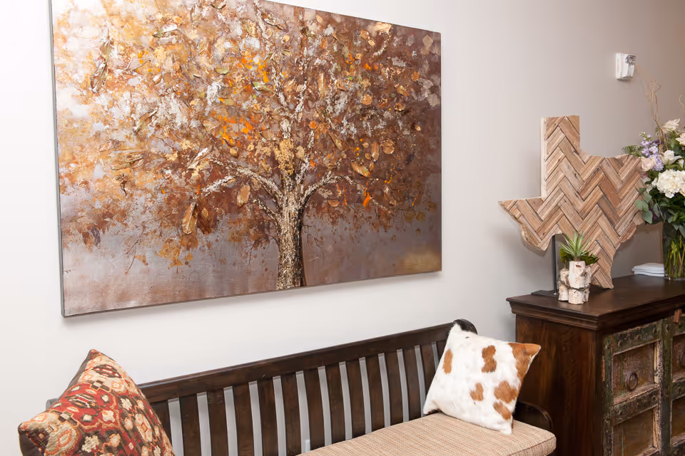 A cozy interior corner featuring a wooden bench with two decorative pillows, one with a red patterned design and the other with a white and brown spotted pattern. Above the bench hangs a large textured painting of a tree with autumn-colored leaves. To the right, there is a wooden cabinet topped with a wooden Texas-shaped decor piece, a small potted plant, and a vase of flowers.