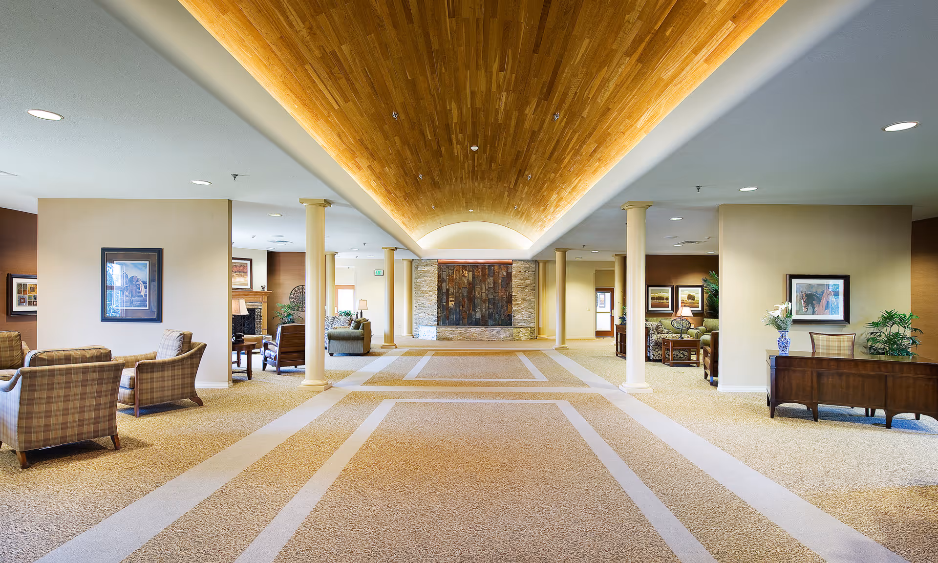 Spacious and well-lit interior common area with a high wooden ceiling, beige carpet with geometric patterns, cream-colored walls, and several seating arrangements including armchairs and sofas. The room features decorative columns, framed artwork on the walls, and plants placed on tables and stands.