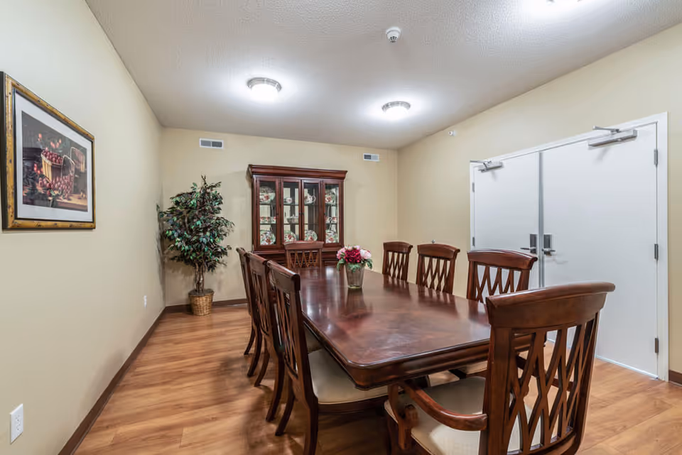 Long wooden dining table with chairs and a floral centerpiece in a private dining room with a china cabinet and double doors.