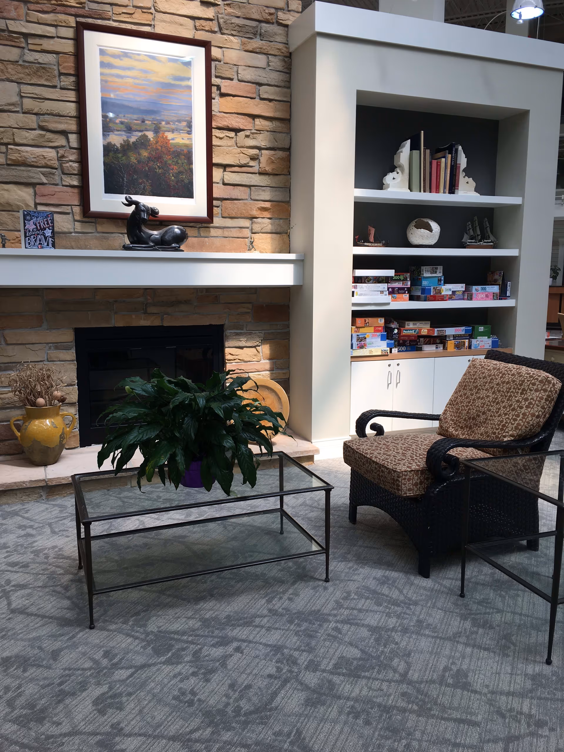 A cozy indoor seating area featuring a cushioned wicker armchair and a glass coffee table with a green potted plant on top. Behind the seating area is a stone fireplace with a white mantel displaying a framed landscape painting, a small sculpture, and a decorative sign. To the right of the fireplace is a built-in white bookshelf filled with books and board games.