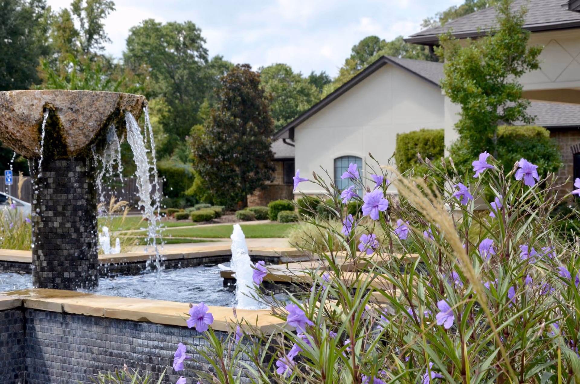 Outdoor view of a landscaped garden area at Providence Park Rehabilitation And Skilled Nursing featuring a stone water fountain with water flowing from the top, surrounded by purple flowers and greenery, with a building in the background.