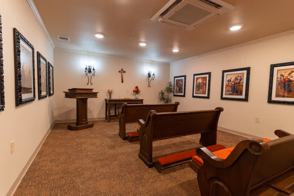 A small chapel room with wooden pews and kneelers, a wooden lectern, and a table with flowers. The walls are decorated with framed religious paintings and a crucifix. The room is softly lit with wall sconces and ceiling lights.